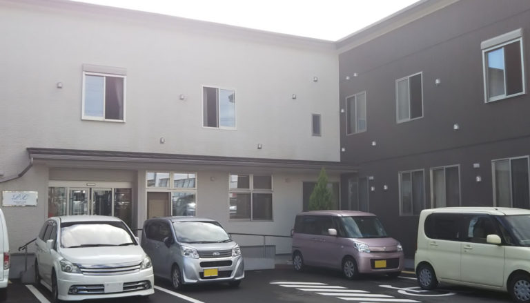 施設の駐車場と建物外観。複数台の車が停めてあり、モダンな2層の建物が見える。